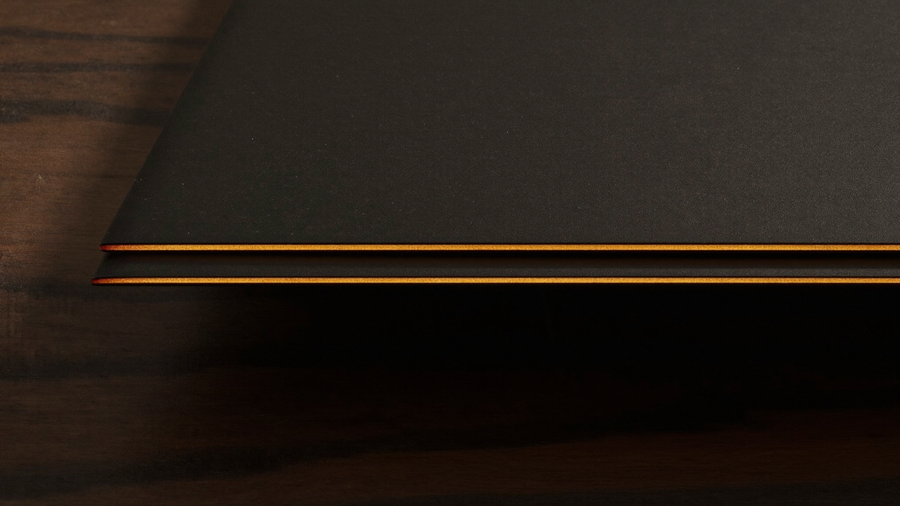 Extreme macro of two razor-thin matte black device edges stacked on dark walnut, each catching a warm amber specular line, illustrating foldable thickness measurement