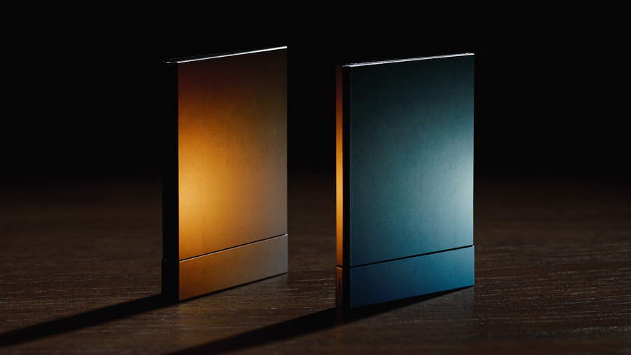 Two closed foldable phone silhouettes standing face to face on dark walnut in a blacked-out studio, the left lit warm amber and the right lit cool steel blue, long shadows stretching toward the viewer